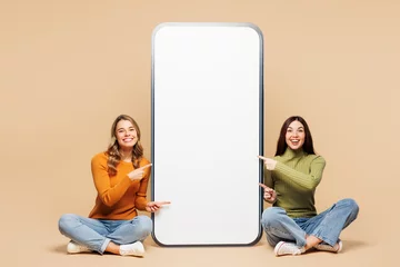 Gordijnen Full body young friends two women wear orange green shirt casual clothes together sit point finger on big huge blank screen mobile cell phone smartphone with area isolated on plain beige background. © ViDi Studio