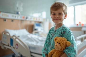 A young boy dressed in hospital clothes stands next to a bed in a hospital ward holding a teddy bear