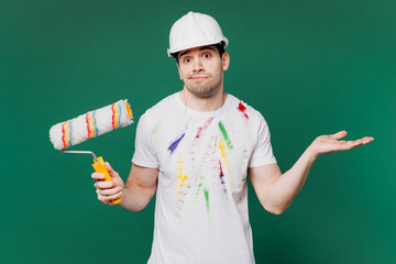 Young employee laborer handyman man wear stained t-shirt hardhat hat hold paint roller spread hands isolated on plain green background. Instruments for renovation apartment room. Repair home concept.