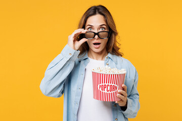 Young surprised shocked sad woman wear blue shirt white t-shirt casual clothes lower 3d glasses watch movie film hold bucket of popcorn in cinema isolated on plain yellow background studio portrait.