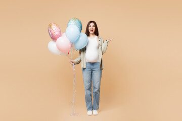 Full body happy young pregnant woman future mom in grey shirt with belly stomach tummy with baby hold bunch pink blue inflated balloons isolated on plain beige background. Maternity pregnancy concept.