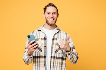 Young fun smiling happy Caucasian man he wear brown shirt casual clothes hold in hand use mobile cell phone show thumb up isolated on plain yellow orange background studio portrait. Lifestyle concept.
