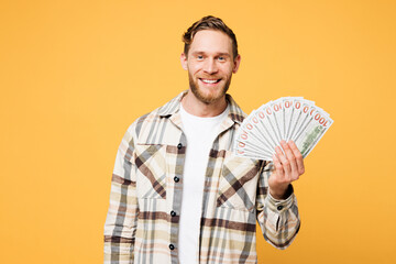 Young smiling happy Caucasian man he wear brown shirt casual clothes hold in hand fan of cash money in dollar banknotes isolated on plain yellow orange background studio portrait. Lifestyle concept.