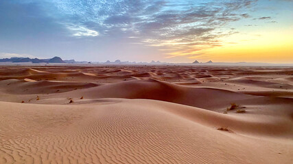Landscape of Erg Admer in the Sahara desert, Algeria. Waves of dunes at sunrise with, in the distance, the rock formations of Tassilis - 750082042