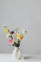 Stylish spring flowers in vase with ribbon on rustic white table. Happy womens day and Mother's day. Beautiful daffodils and tulips gentle bouquet still life. Space for text