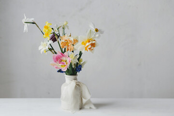 Stylish spring flowers in vase with ribbon on rustic white table. Happy womens day and Mother's day. Beautiful daffodils and tulips gentle bouquet still life. Space for text