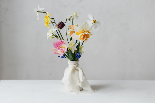 Stylish spring flowers in vase with ribbon still life. Beautiful daffodils and tulips gentle bouquet on rustic white table. Space for text. Happy womens day and Mother's day