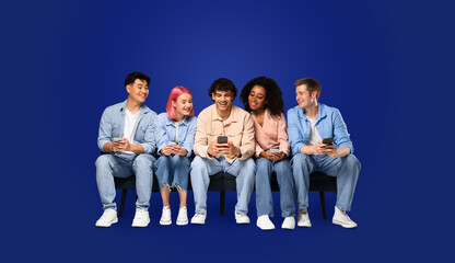 A group of five young, multicultural friends sitting on a bench, engaged with their smartphones