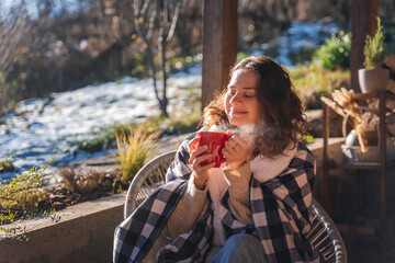 Young woman enjoying a sunny early spring morning on the terrace, wrapping herself in a blanket and drinking hot drink - 750089881