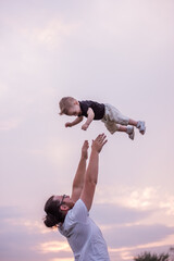 Young, diversified father throws little son into air against backdrop of soft pastel sky at sunset....
