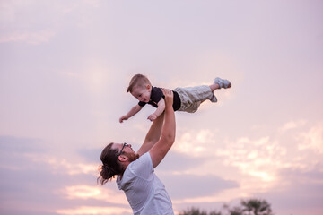 Young, diversified father throws little son into air against backdrop of soft pastel sky at sunset. Toddler is gleefully thrown in the air by parent, with background dusk, symbolizing joy and freedom