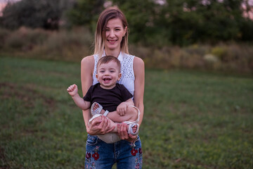 Fototapeta premium Young woman smiles, holds toddler son in her arms, standing in lush field with the soft glow of evening light. The concept of tactility, maternal connection with the child.