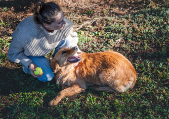 Young happy charming girl playing with her retriever dog with a ball on green grass. - 750090608