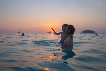 A father carries his child to play in sea water by the beach at sunset. .The father is taking the opportunity to bond with his child and enjoy a peaceful evening. .The sun is on the palm of his hand.