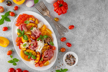 Caprese salad with mozzarella, tomatoes, prosciutto and grilled peaches on a white plate on a gray concrete background. Fresh summer salad. Top view.