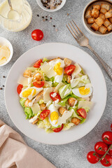 Caesar salad with lettuce, grilled chicken breast, boiled eggs, tomatoes, parmesan cheese and croutons in a ceramic plate on a concrete background. Top view.