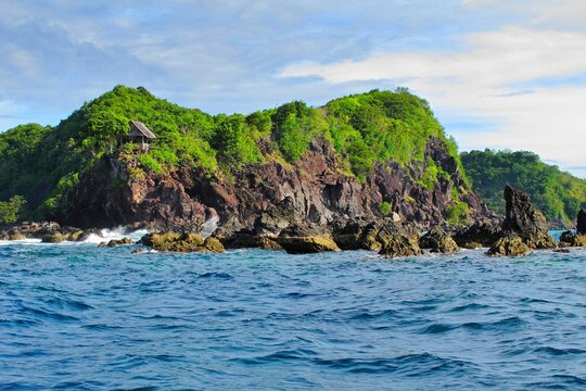 Vivid green tropical island with small house, dangerous rocks and waves on the ocean. View on the romantic dangerous rocky shoreline, travel picture.