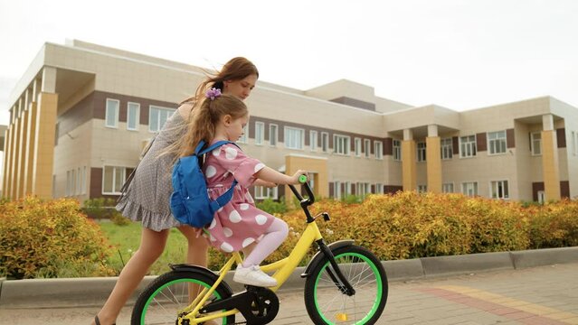 Cute little baby girl pupil with backpack riding on bike at autumn schoolyard before lesson with mother. Happy family playful female kid daughter and mom spending time together driving cycle outdoor