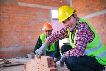 Construction worker bricklaying wall in a home development real estate, happy smiling young Asian male builder doing labor work building a house. Working with a co-worker in the background.