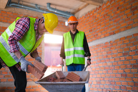 Construction worker bricklaying carrying bricks into wheelbarrow, happy smiling young Asian male builder doing labor work building a house. Working with a co-worker in the background.