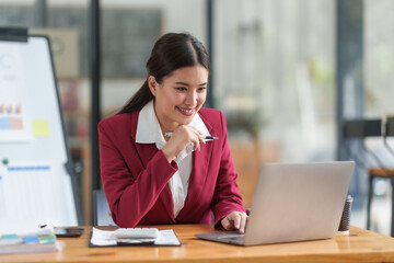 Fototapeta premium Cheerful asian businesswoman work diligently on her laptop in modern office.