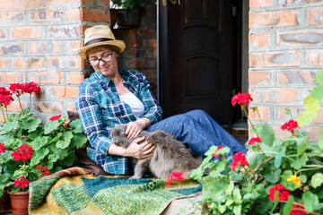 Happy woman and cat relaxing together on cozy home terrace in summer outdoors. Middle-aged farmer...