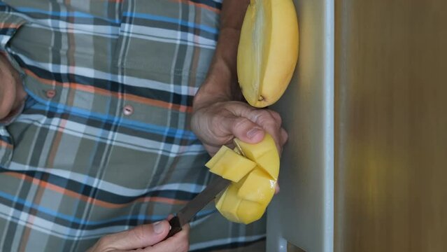 Vertical video. Close-up of man cutting resh ripe mango fruit with knife indoors, slow motion