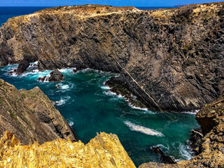 The image shows a rugged coastal landscape with steep cliffs surrounding a small inlet of turquoise sea. The sea is turbulent with white waves crashing against the rocky shoreline.