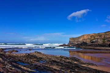Waves hit the rocky shore with layered rock formations under the clear sky and beside a large cliff.