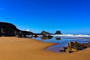 A sandy beach with rocky outcrops, waves gently crashing, under a clear blue sky.