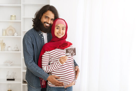 Pregnant Muslim Couple Holding Baby Ultrasound Scan While Standing In Home Interior