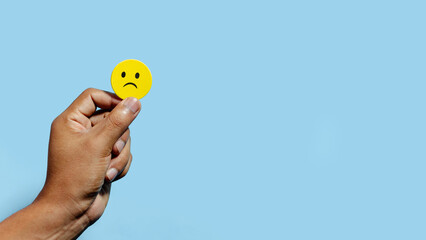 hand holding a  sad emoticon against a light blue background in the foreground. copyspace