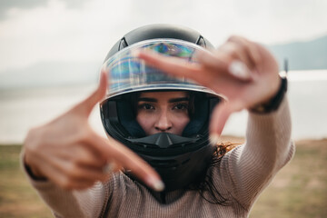 A creative shot of a Latina woman framing the camera with her hands while wearing a motorcycle helmet, with a lake backdrop