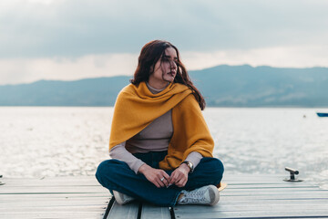 A young woman sits cross-legged on a wooden dock, wrapped in a yellow shawl, looking pensively across the waters of a mountain lake - Powered by Adobe