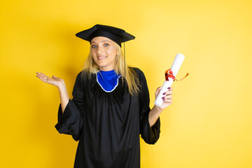 Beautiful blonde young woman wearing graduation cap and ceremony robe clueless and confused...