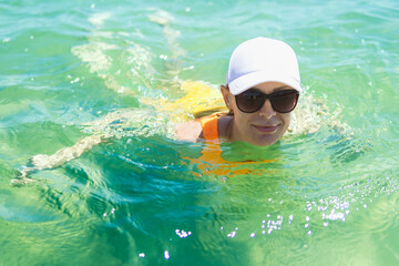 Fototapeta na wymiar A happy woman in a swimsuit, sunglasses, and white cap enjoys a swim in the sea during summer, capturing the essence of carefree oceanic bliss.