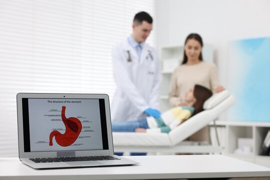Gastroenterologist examining girl in clinic, focus on laptop with image of stomach on white table