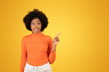 An inquisitive young woman with afro hair points upwards while wearing a bright orange sweater
