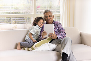 Positive Indian granddad showing online learning application to little grandkid holding tablet,...