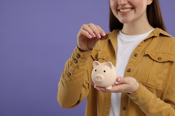 Woman putting coin into piggy bank on purple background, closeup. Space for text