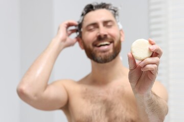 Happy man showing solid shampoo bar in bathroom, selective focus
