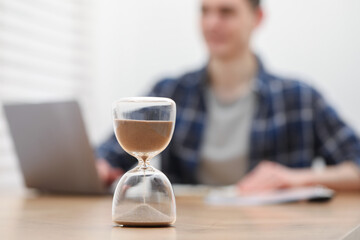 Hourglass with flowing sand on desk. Man taking notes while using laptop indoors, selective focus