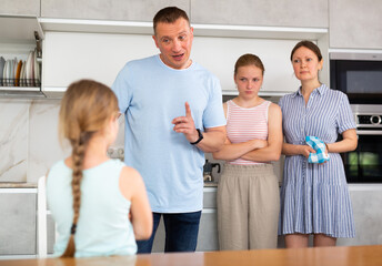 Family scene in kitchen, unpleasant conversation. Girl with hair in braids and crossed arms on chest stands in front of parents and listens to instructions about her bad behavior