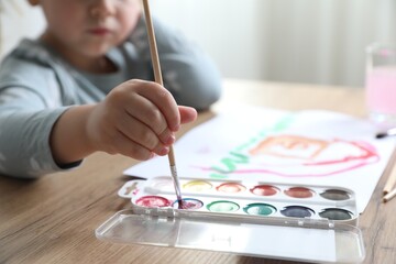 Little girl with brush drawing at wooden table indoors, selective focus. Child`s art