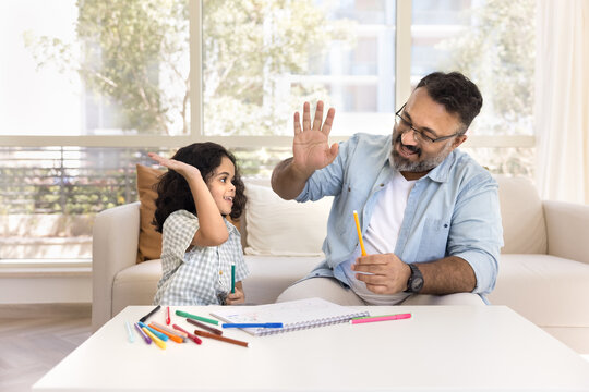 Happy Indian grandpa and sweet preschool granddaughter giving high five over paper album drawing colored pencils, smiling, enjoying creative achievement, success in artistic hobby