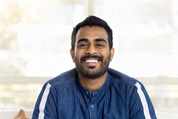Obraz premium Cheerful attractive young Indian man head shot front portrait. Happy handsome guy in casual denim shirt looking at camera with perfect toothy smile, showing white teeth. Video call screen
