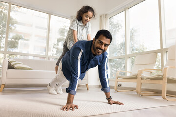 Happy strong athletic Indian dad piggybacking little daughter, keeping static yoga plank on floor,...