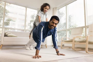 Foto op Canvas Happy strong athletic Indian dad piggybacking little daughter, keeping static yoga plank on floor, doing pushups with kids weight, looking at camera, exercising for strength, training body © fizkes