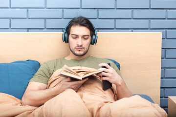 Obraz premium Young man lying at the bed, reading a book and listening audiobook at the same time