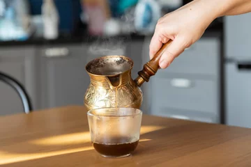 Fotobehang A woman pouring coffee into a cup from a copper Turkish coffee pot © Vitaliy